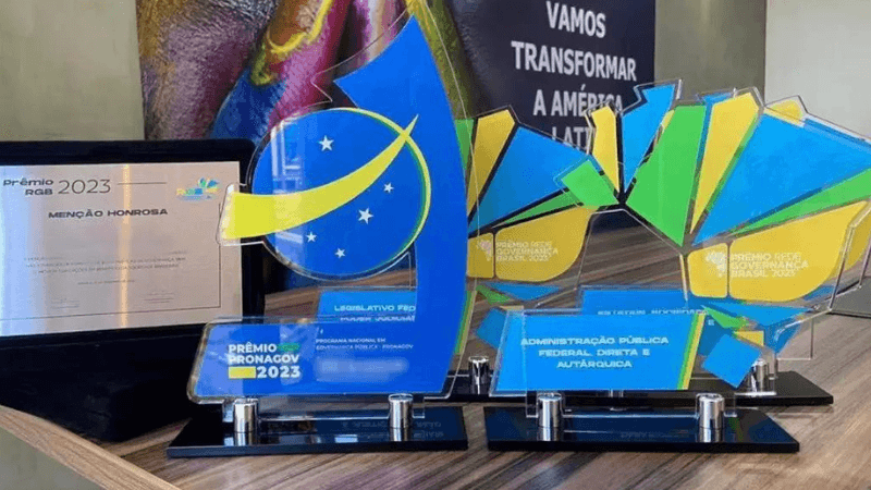 três troféus e uma placa de homenagem estão sobre uma mesa para divulgar premiação nacional